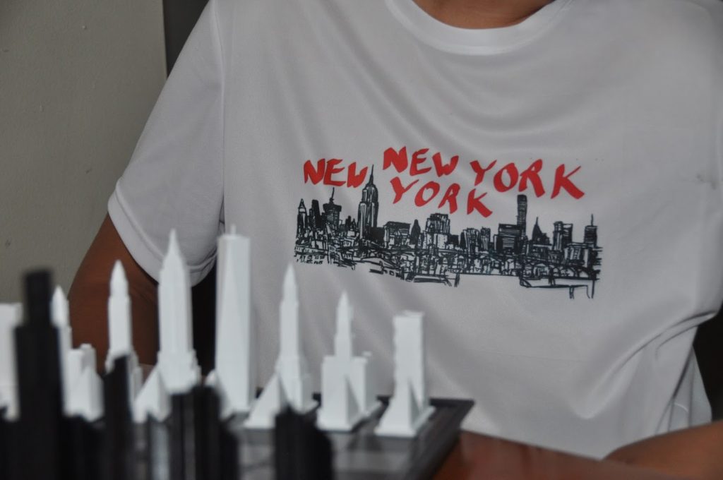Displayed is a white shirt with a dye-sublimated design that states "New York" twice with a vector picture of the city skyline. 