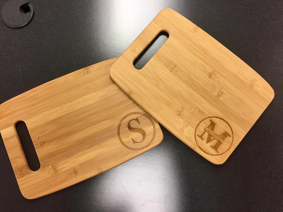 Two laser engraved cutting boards. One lays slightly on the other. One cutting board, on left, has a circular design with an S initial. The other displays a circular design with an M initial. 