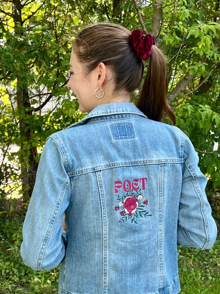 A young female wears a blue jean jacket. The back of the jean jacket features a floral design with the word poet" displayed above. 