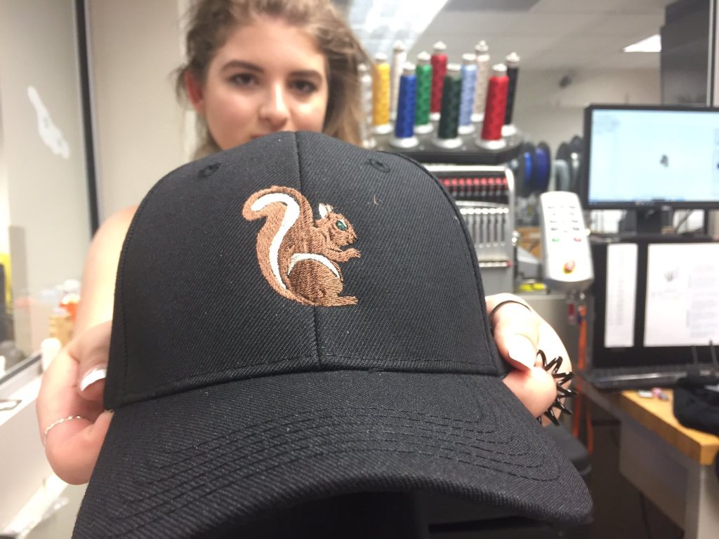 Young female student holds out a black embroidered cap with a brown squirrel design. 