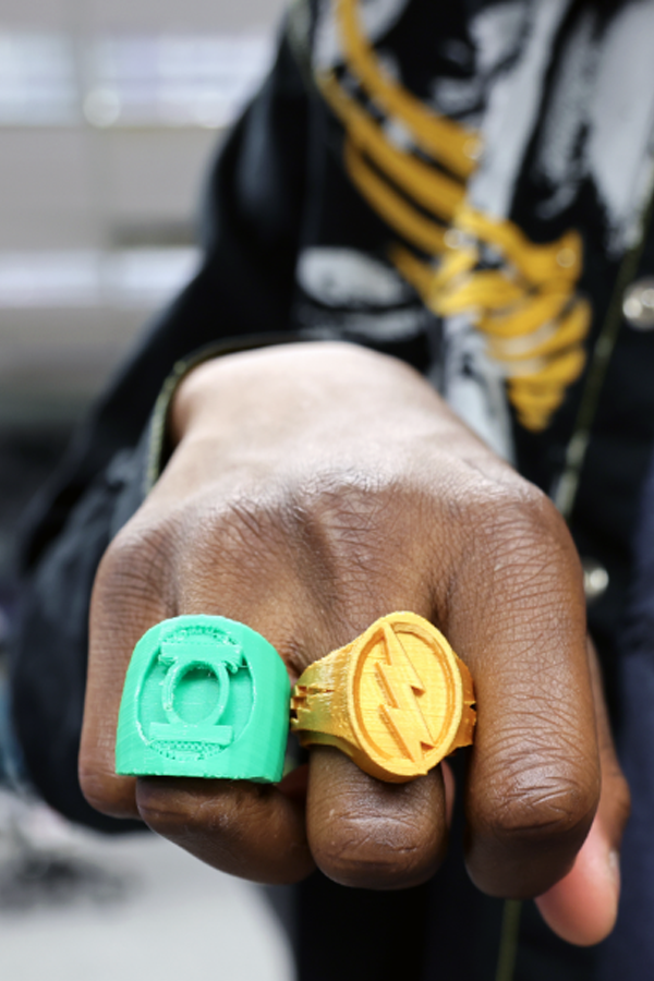 Two 3D printed rings displayed on students hand in fist. One ring is green with a design and the other ring is gold with a lightning bolt design.
