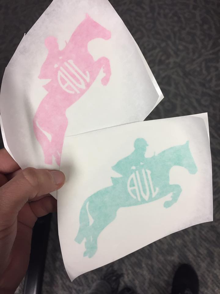 Two vinyl cut stickers are held. The design is of a person on a horse with the initials "AVL". The top sticker is pink and the bottom sticker is green. 